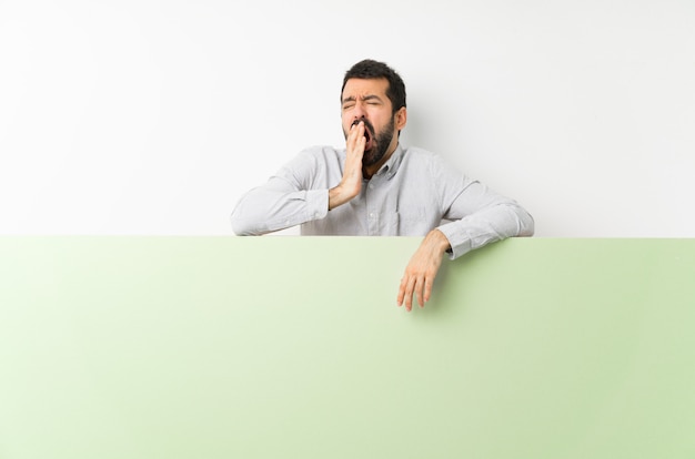 Joven guapo con barba sosteniendo una gran pancarta vacía verde bostezando y cubriendo la boca abierta con la mano