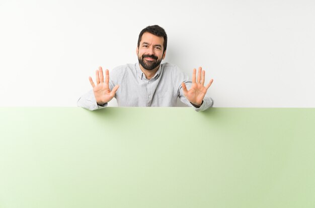 Joven guapo con barba sosteniendo una gran pancarta vacía verde contando diez con los dedos