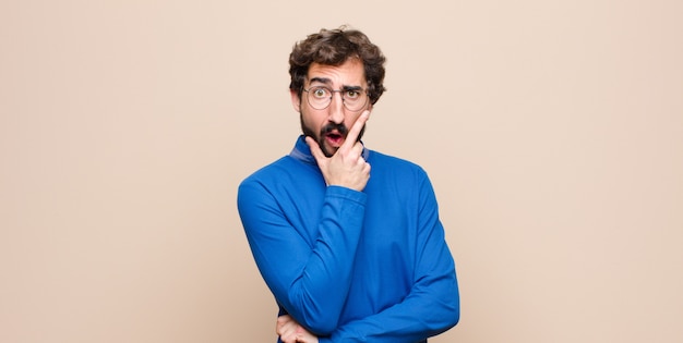 Joven guapo con la boca y los ojos bien abiertos y la mano en la barbilla, sintiéndose desagradablemente sorprendido, diciendo qué o wow contra la pared