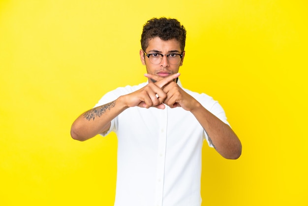 Joven guapo brasileño aislado sobre fondo amarillo haciendo gesto de parada con la mano para detener un acto