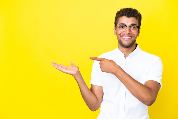 Joven guapo brasileño aislado sobre fondo amarillo sosteniendo copyspace imaginario en la palma para insertar un anuncio