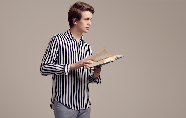 Joven guapo en camisa a rayas leyendo un libro sobre fondo gris