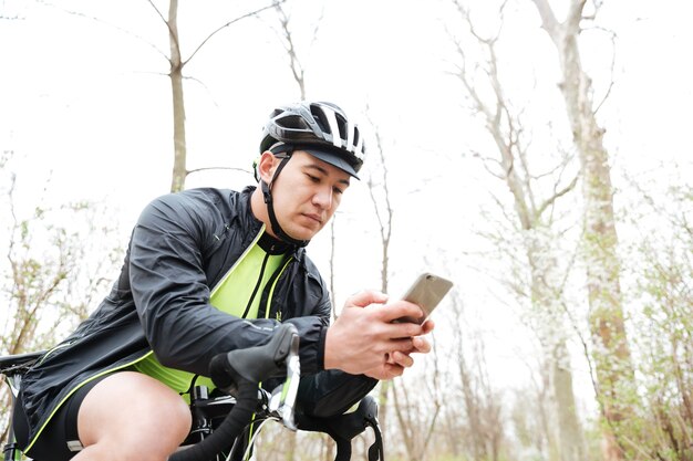 Joven guapo en casco de ciclismo con bicicleta con smartphone en el parque