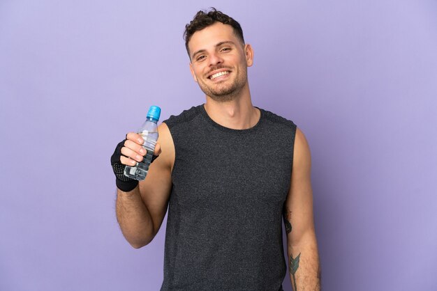 Joven guapo caucásico aislado en morado con botella de agua deportiva