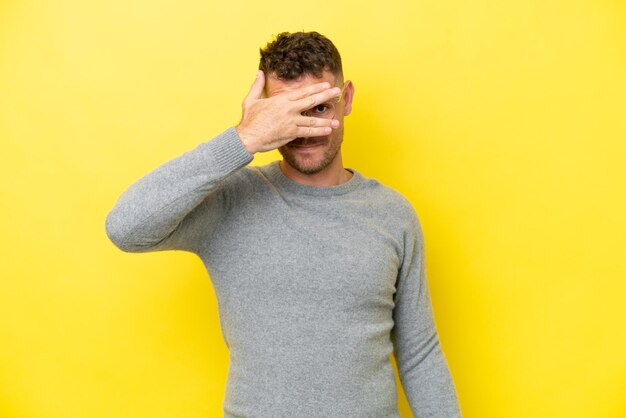 Joven guapo caucásico aislado sobre fondo amarillo cubriendo los ojos con las manos y sonriendo