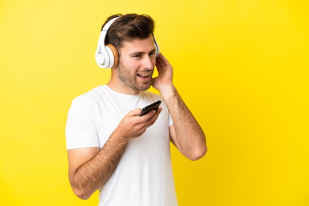 Joven guapo caucásico aislado sobre fondo amarillo escuchando música con un móvil y cantando