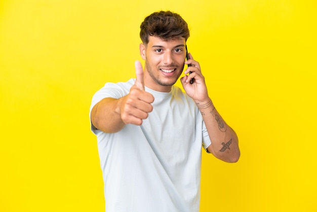 Joven guapo caucásico aislado sobre fondo amarillo manteniendo una conversación con el móvil mientras hace los pulgares para arriba