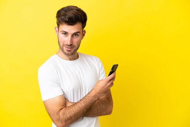 Joven guapo caucásico aislado sobre fondo amarillo sosteniendo un teléfono móvil y con los brazos cruzados.