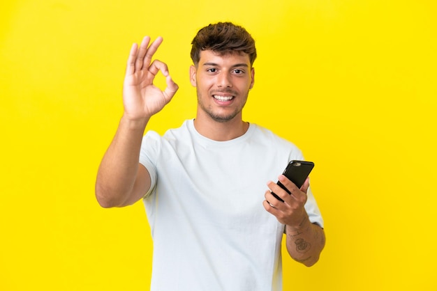 Joven guapo caucásico aislado sobre fondo amarillo con teléfono móvil y haciendo el signo de OK