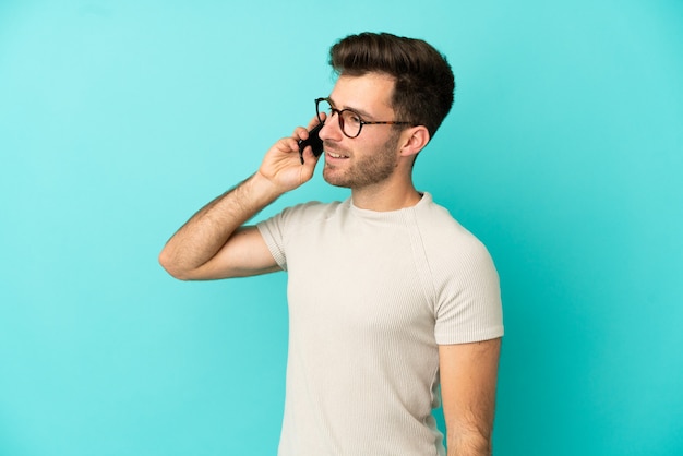 Joven guapo caucásico aislado sobre fondo azul manteniendo una conversación con el teléfono móvil