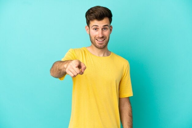 Joven guapo caucásico aislado sobre fondo azul sorprendido y apuntando hacia el frente