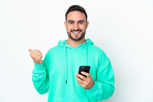 Joven guapo caucásico aislado sobre fondo blanco con teléfono móvil y apuntando hacia el lateral