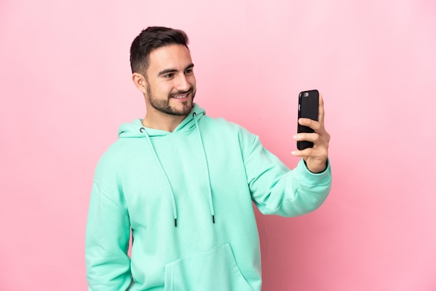 Joven guapo caucásico aislado sobre fondo rosa haciendo un selfie