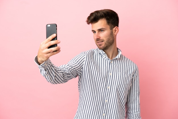 Joven guapo caucásico aislado sobre fondo rosa haciendo un selfie
