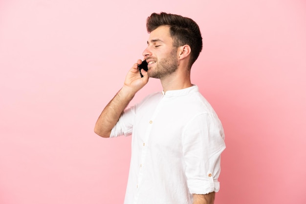 Joven guapo caucásico aislado sobre fondo rosa manteniendo una conversación con el teléfono móvil