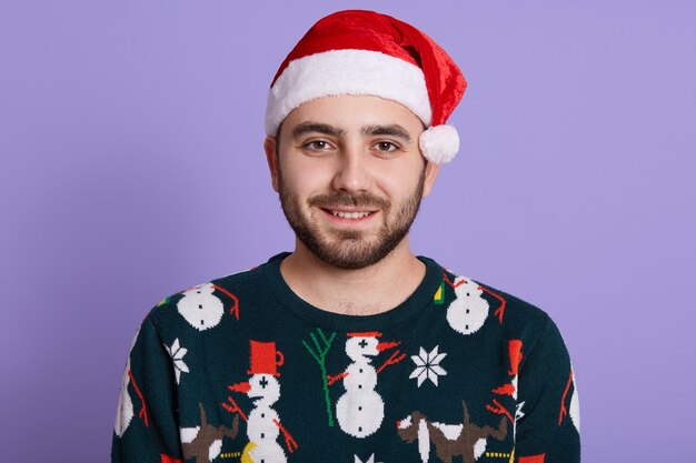 Joven guapo caucásico con gorro de Papá Noel rojo y suéter cálido con estampado divertido se encuentra en el espacio lila