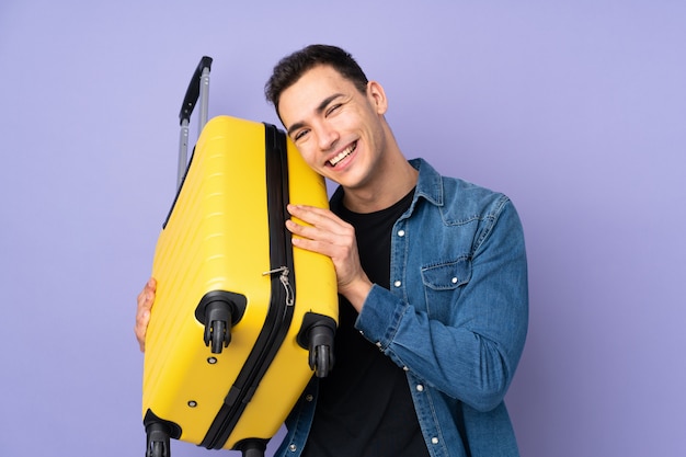 Joven guapo caucásico en pared púrpura en vacaciones con maleta de viaje