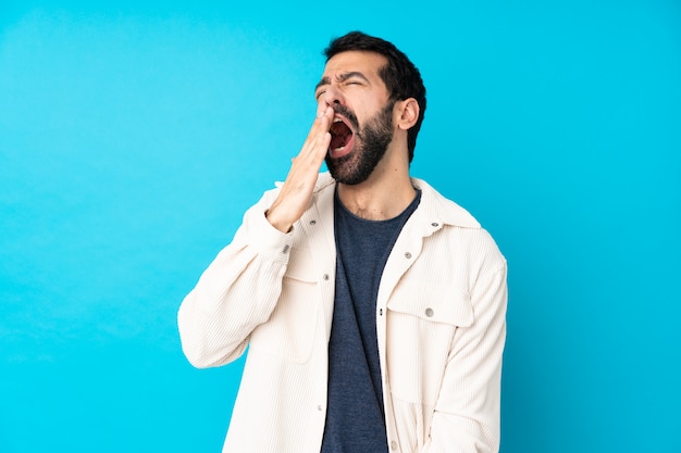 Foto joven guapo con chaqueta de pana blanca sobre pared azul aislada bostezando y cubriendo la boca abierta con la mano