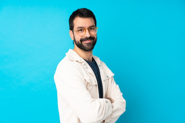 Joven guapo con chaqueta de pana blanca sobre pared azul aislada con gafas y sonriendo