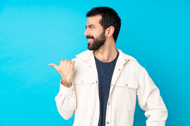 Joven guapo con chaqueta de pana blanca sobre pared azul aislado apuntando hacia un lado para presentar un producto