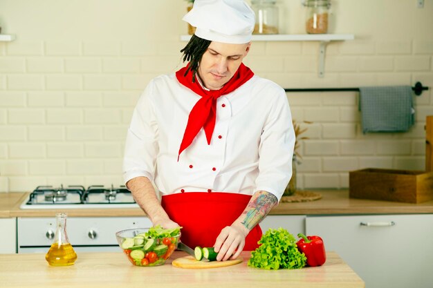 Joven guapo chef cortando pepino verde sobre tabla de madera en la cocina Chico positivo con rastas en uniforme preparando ensalada de verduras