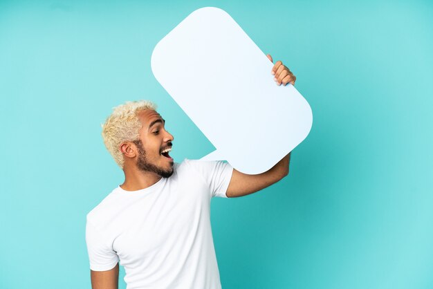 Joven guapo colombiano aislado sobre fondo azul sosteniendo un bocadillo de diálogo vacío