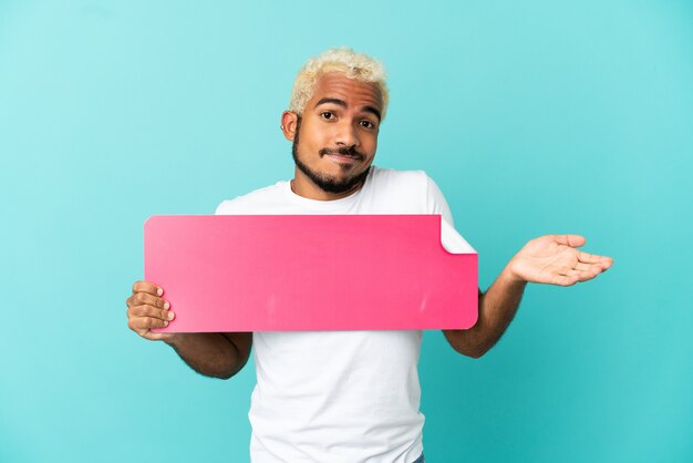 Joven guapo colombiano aislado sobre fondo azul sosteniendo un cartel vacío y tener dudas