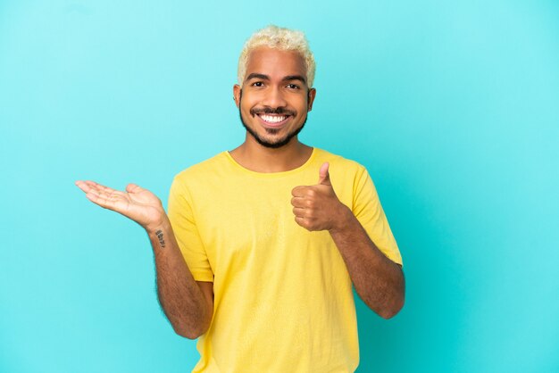 Joven guapo colombiano aislado sobre fondo azul sosteniendo copyspace imaginario en la palma para insertar un anuncio y con los pulgares hacia arriba