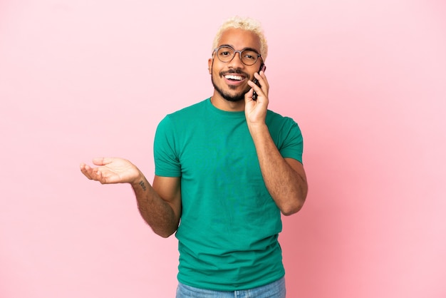 Joven guapo colombiano aislado sobre fondo rosa manteniendo una conversación con el teléfono móvil con alguien