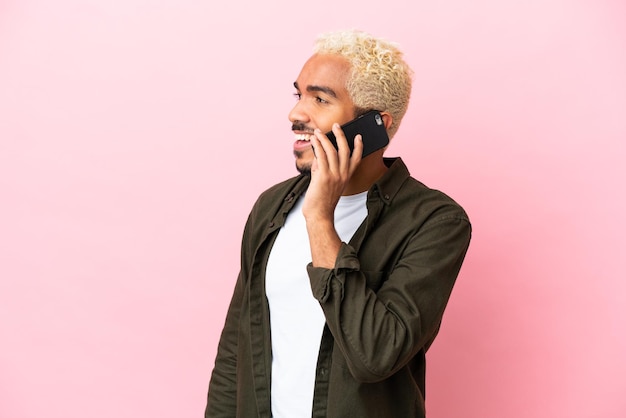 Joven guapo colombiano aislado sobre fondo rosa manteniendo una conversación con el teléfono móvil con alguien