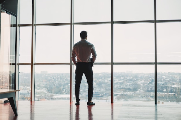 Un joven guapo con una computadora portátil de pie en el fondo de una gran ventana panorámica en el cielo una oficina moderna con grandes ventanas y un trabajador de oficina