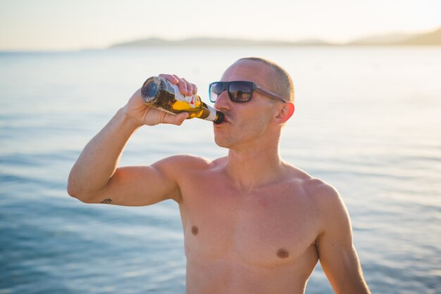 joven guapo deportivo bebiendo cerveza en la playa puesta de sol