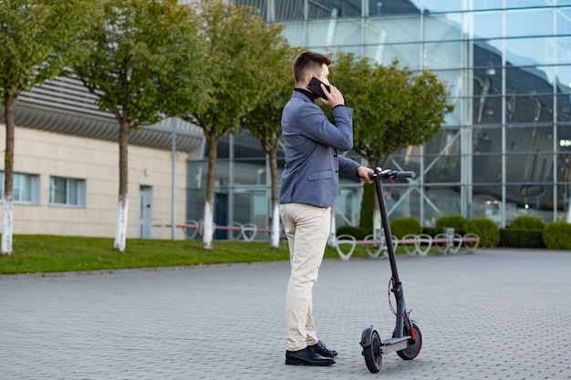 Joven guapo con e-scooter de pie en la acera cerca del aeropuerto en la puesta de sol y hablando por el teléfono inteligente. Transporte urbano de moda en scooter eléctrico moderno. Concepto de movilidad ecológica