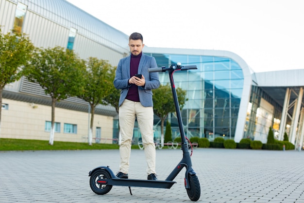 Joven guapo con e-scooter de pie en la acera cerca del aeropuerto en la puesta de sol y hablando por el teléfono inteligente