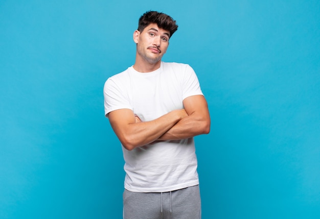 Joven guapo encogiéndose de hombros, sintiéndose confundido e inseguro, dudando con los brazos cruzados y la mirada perpleja