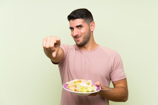 Joven guapo con ensalada en la pared verde te señala con una expresión segura