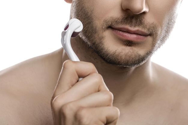 Joven guapo está usando derma roller para el crecimiento de la barba sobre fondo blanco.