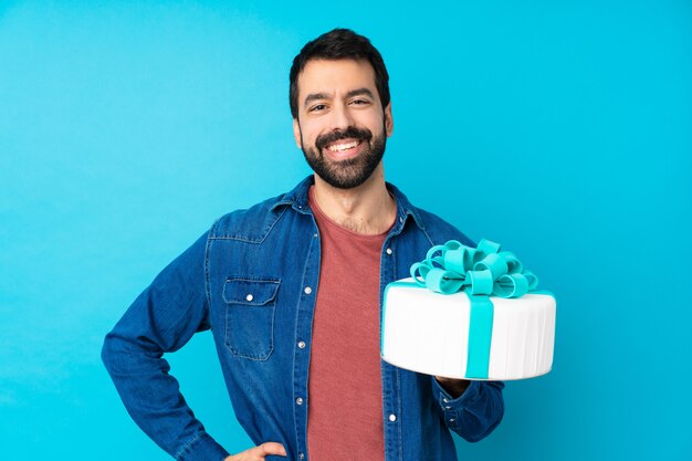 Joven guapo con un gran pastel sobre azul posando con los brazos en la cadera y sonriendo