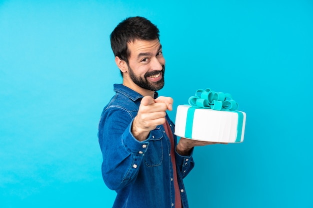 Joven guapo con un gran pastel sobre la pared azul aislada te señala con el dedo mientras sonríe