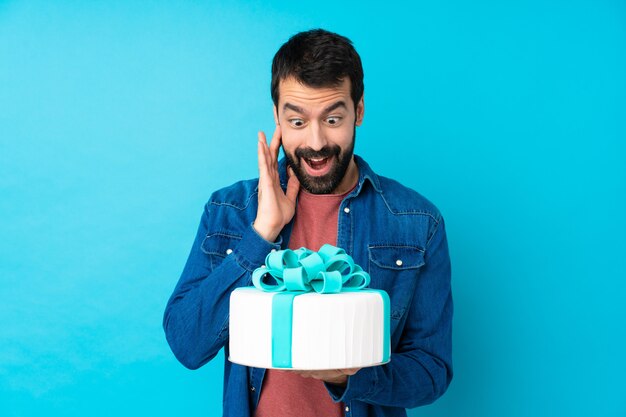 Foto joven guapo con un gran pastel sobre pared azul aislado con sorpresa y expresión facial conmocionada