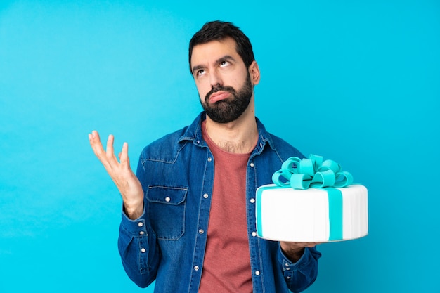 Joven guapo con un gran pastel sobre pared azul frustrado por una mala situación