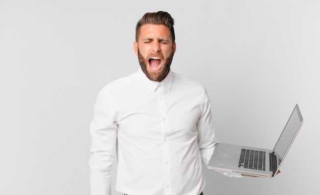 Joven guapo gritando agresivamente, luciendo muy enojado y sosteniendo una computadora portátil