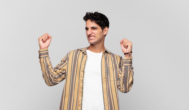 Joven guapo gritando triunfalmente, con aspecto de ganador emocionado, feliz y sorprendido, celebrando