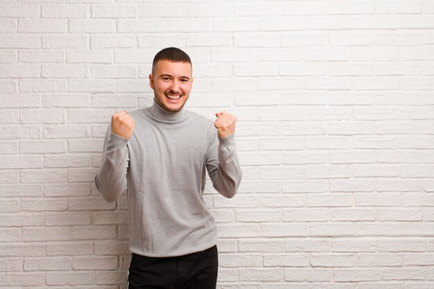 Joven guapo gritando triunfalmente, riendo y sintiéndose feliz y emocionado mientras celebra el éxito contra la pared plana