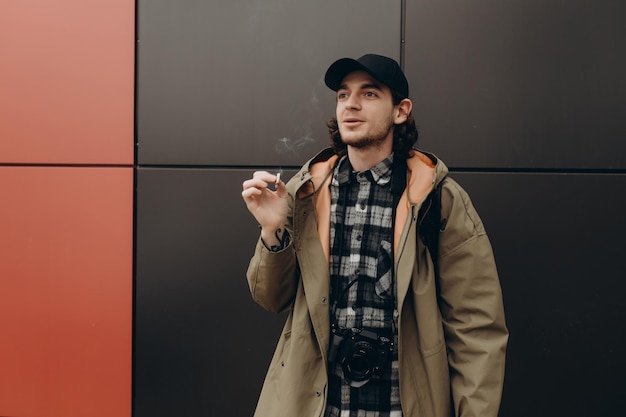 Joven guapo hipster barbudo fumando cigarrillo cerca de la pared en la calle