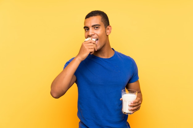 Joven guapo hombre morena sobre pared amarilla desayunando leche y sosteniendo pastel de muffin