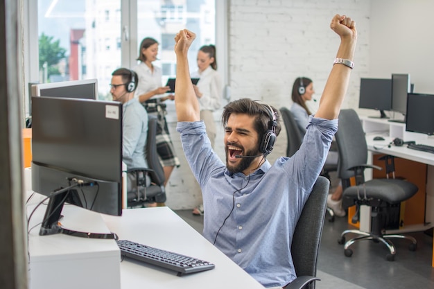 Joven y guapo hombre de negocios barbudo celebrando el éxito corporativo con los puños levantados en el aire