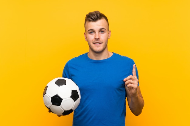 Joven y guapo hombre rubio sosteniendo una pelota de fútbol sobre fondo amarillo aislado con la intención de darse cuenta de la solución mientras levanta un dedo