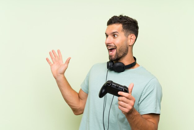 Joven guapo jugando con un controlador de videojuego sobre pared verde aislado con expresión facial sorpresa