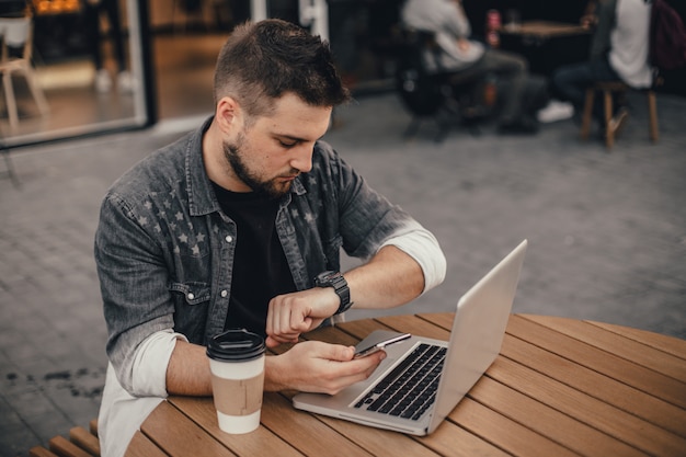 Joven guapo con laptop y taza de café sentarse en la calle de la ciudad